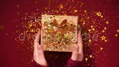 女人手打开礼盒。 闪耀着金色的星星，闪闪发光的纸屑。 在红色背景下打开包装礼物